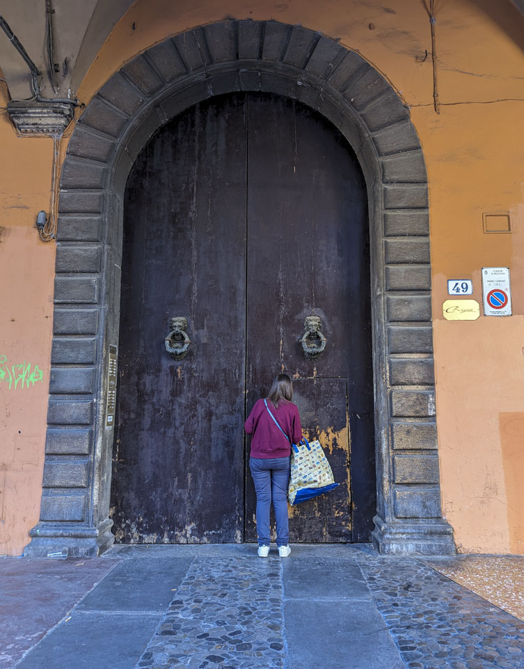 A young woman unlocking a normally sized door that is cut into very large doors that are in an arched frame. The doors are wood, and the frame is stone. The building walls are an orange color.