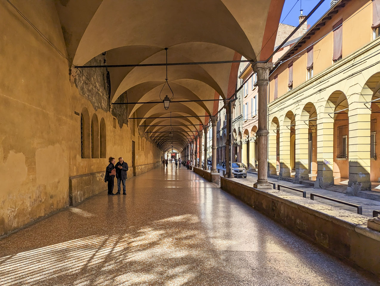 A wide covered sidewalk with a few people on it, and a second covered walkway seen on the other side of the narrow street.