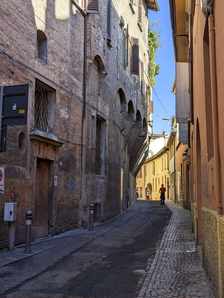 A small street in Bologna with a stone building on the left side that has wider upper floors than the ground floor.