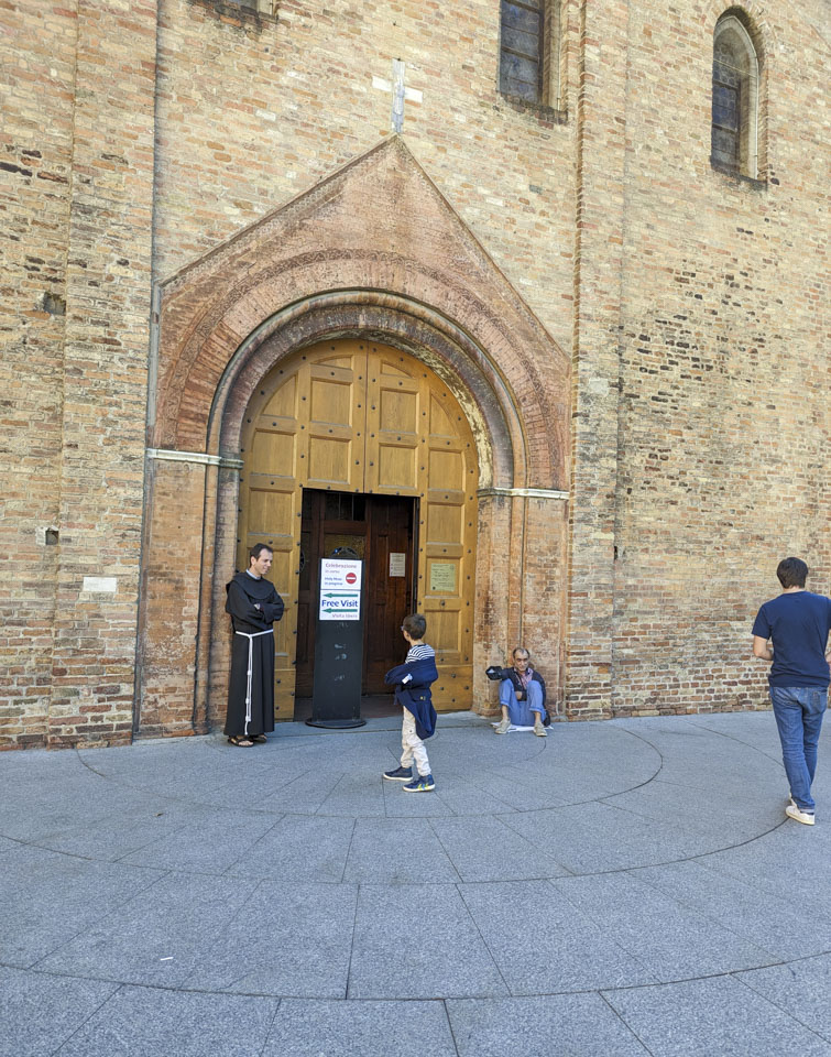 A sign is in front of the church stating that mass is in session; a man in a monk robe is standing next to it, and someone that appears to be a beggar is sitting at the opposite corner of the doorway.