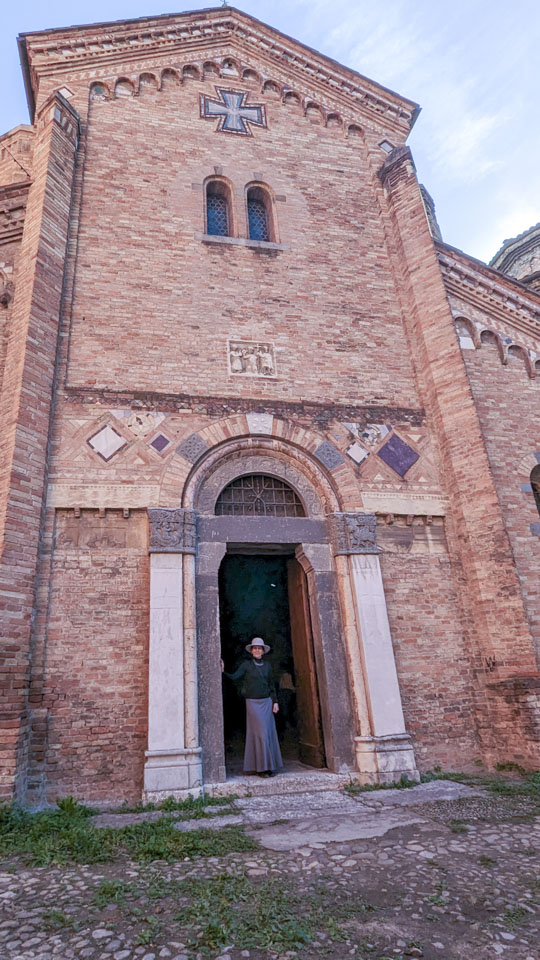 Anne standing in the large doorway into the stone church, which rises high above her. Above the door is a relief of three small figures, and above that is a cross.