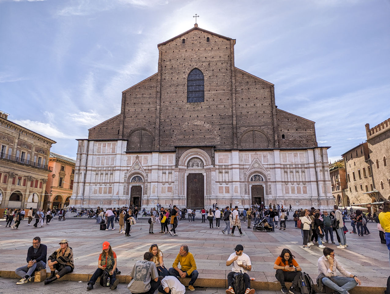 A view of the Basilica from the plaza in front of it. The bottom section has a very finished look, and the top section is simply brick. There is a cross on the top, with the sun behind it.