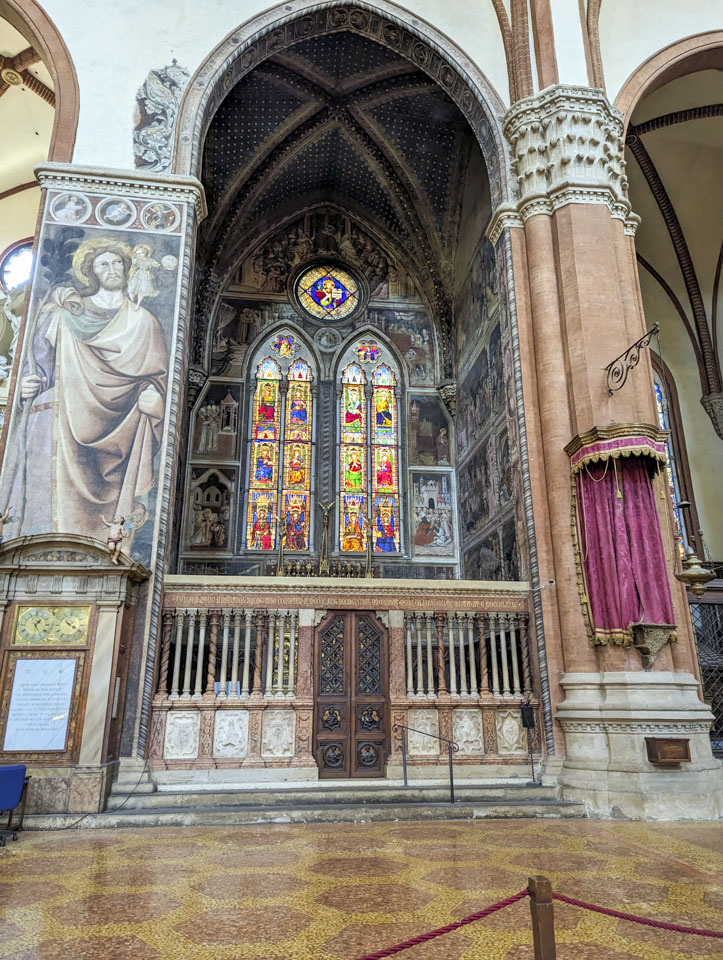 One of the side chapels. There are two tall narrow stained-glass windows with a round one above them. Many small scenes are painted on the walls. 