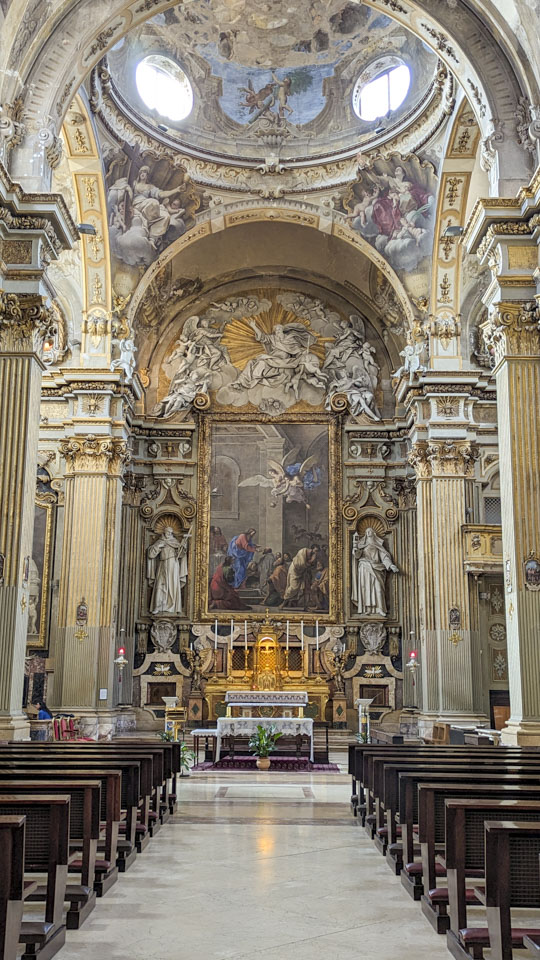 The altar area of a church, with statues of saints and angels, gold painted trim, and a ceiling with figures painted on it.