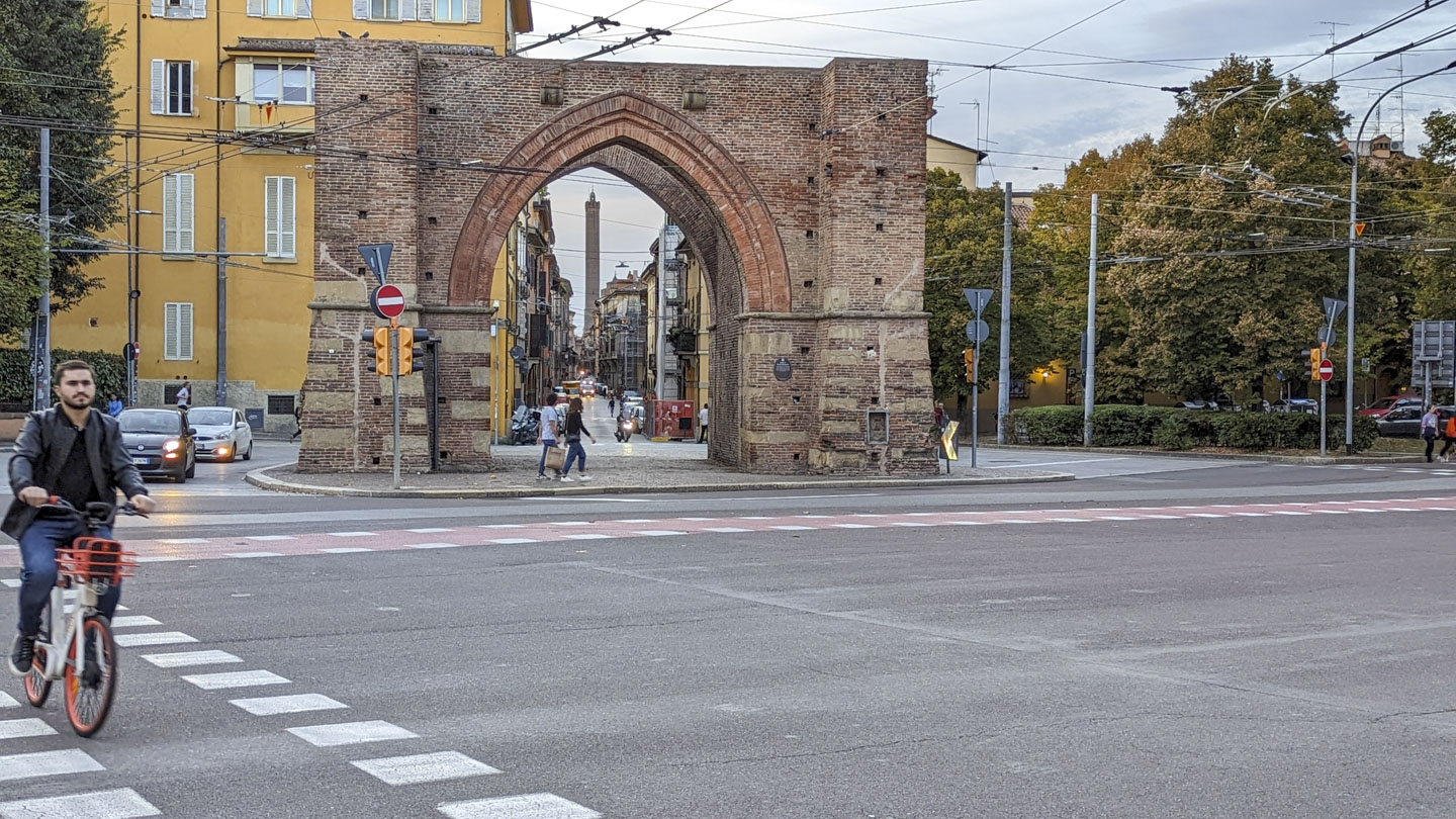 A traffic circle with the remains of a wall in the middle. Through the archway in the wall a tall tower can be seen in the distance.