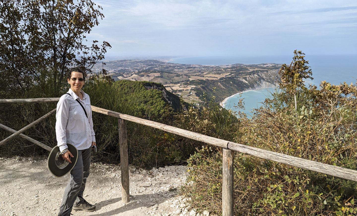 Anne standing next to a minimalist wood fence. In the distance and far below can be seen agricultural land and beyond that are buildings. The land has a steep cliffside down to the water.