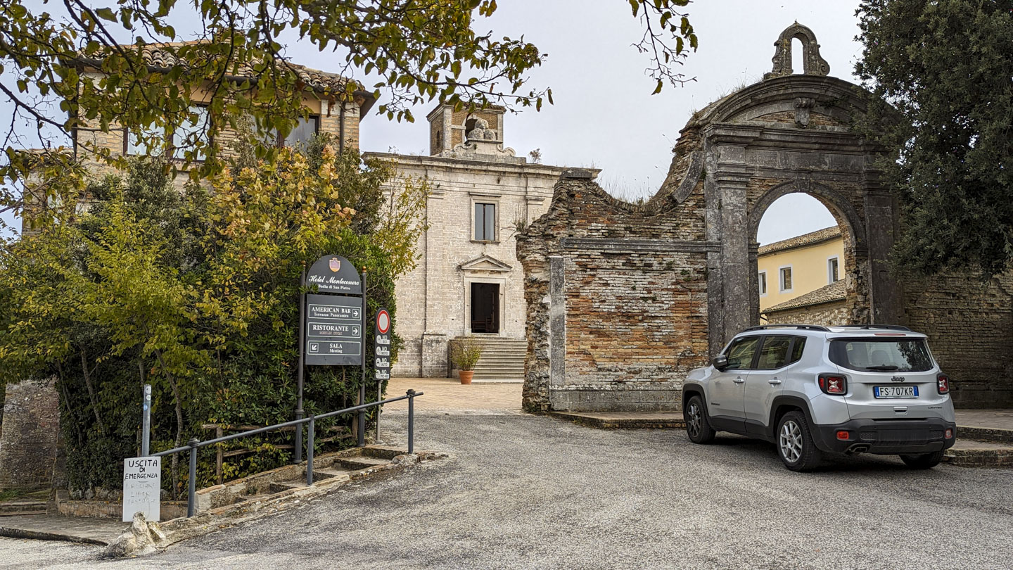 A vehicle is parked in front of an old stone wall that has an arched entrance. Above the entrance is a small arch that might have once held a bell. The wall has a large section missing on the left side, through which a courtyard can be seen and the entrance to a blocky looking church.