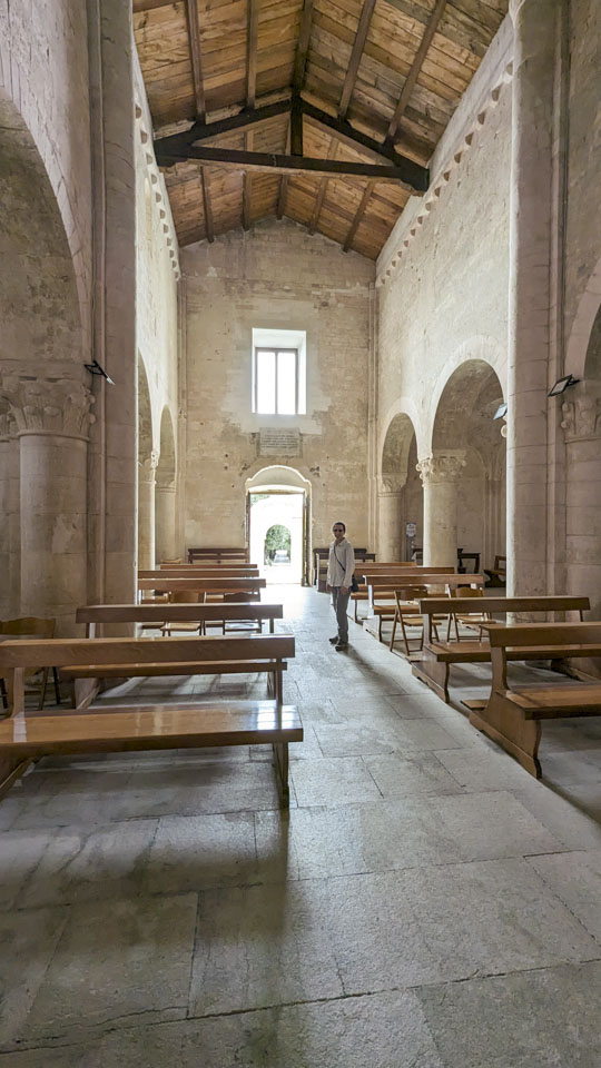 Anne standing in the aisle of an old church that has stone walls and a wooden roof. There are polished wood pews for seating.