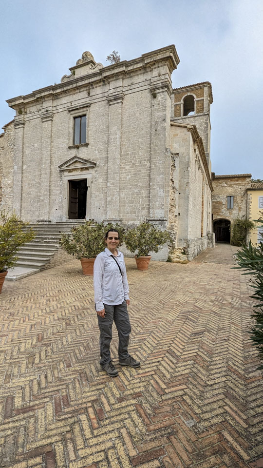 Anne standing in front of the old stone church. It looks like the facade hides a sloped room with a bell tower at the back.