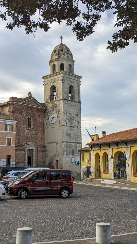 In the middle of the photo is a bell tower with a clock. Attached to it on the left is a red stone front church. A road is in front of it, across which is an orange 1-story building.