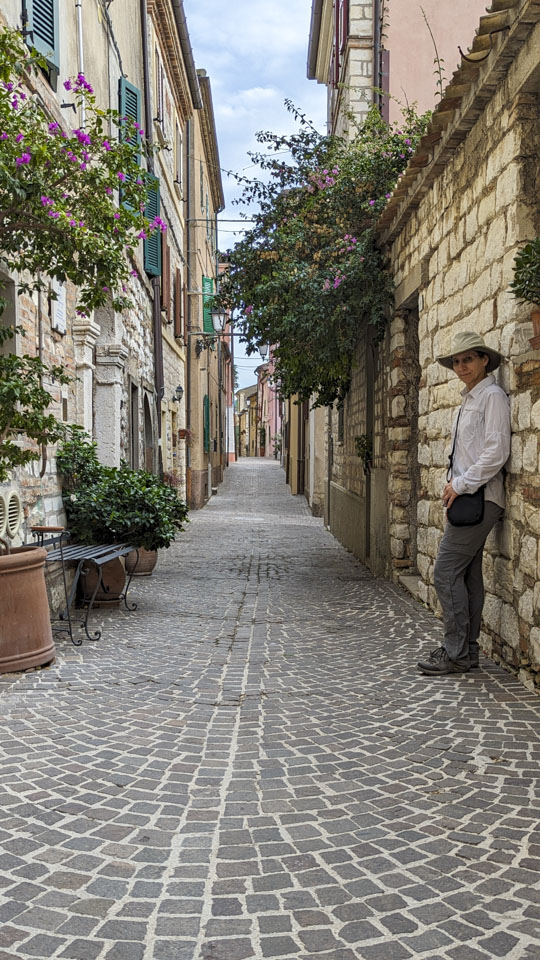 Anne is leaning against a stone wall in a narrow street with buildings on both sides. There are some plants coming over a lower wall behind Anne.