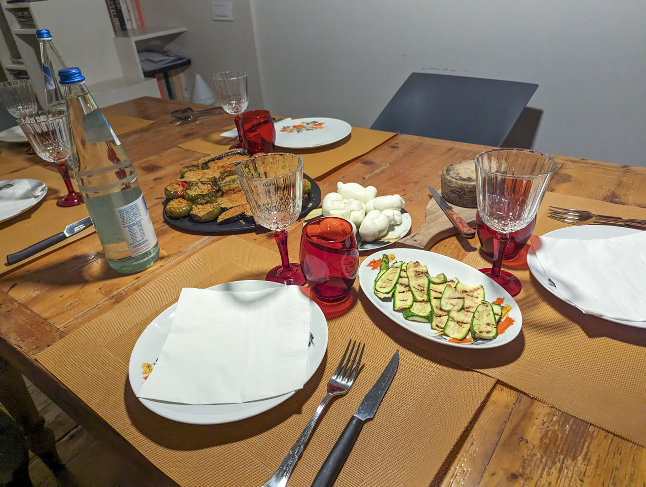 A wooden table with empty plates, glasses, and a couple of bottles of water. There are plates of food as described above, waiting to be served.