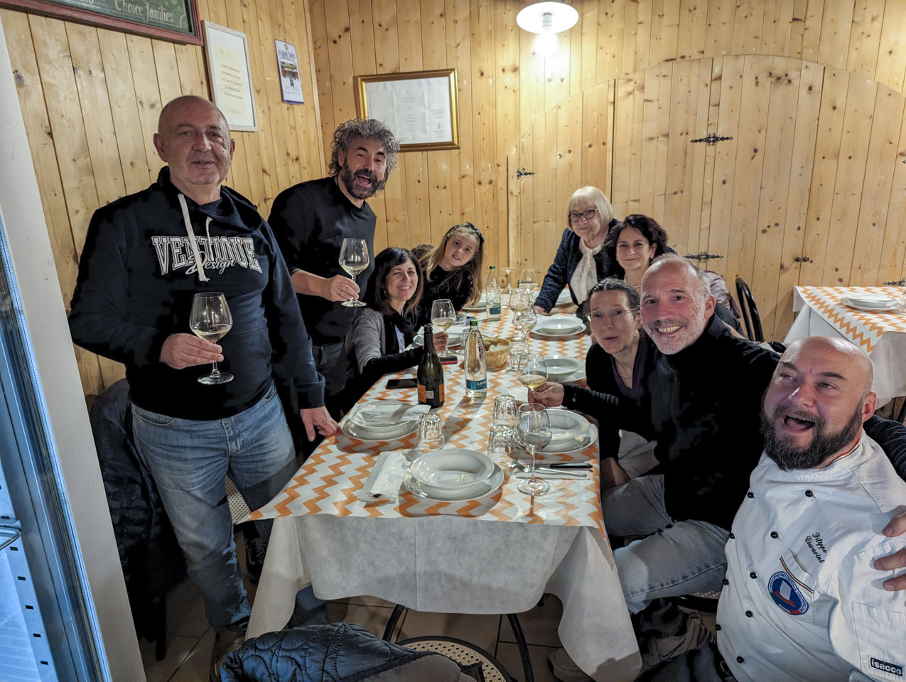 People gathered around a table and looking at the photo taker. There are glasses with some white wine and white empty dishes on the table. The room has light wood walls.