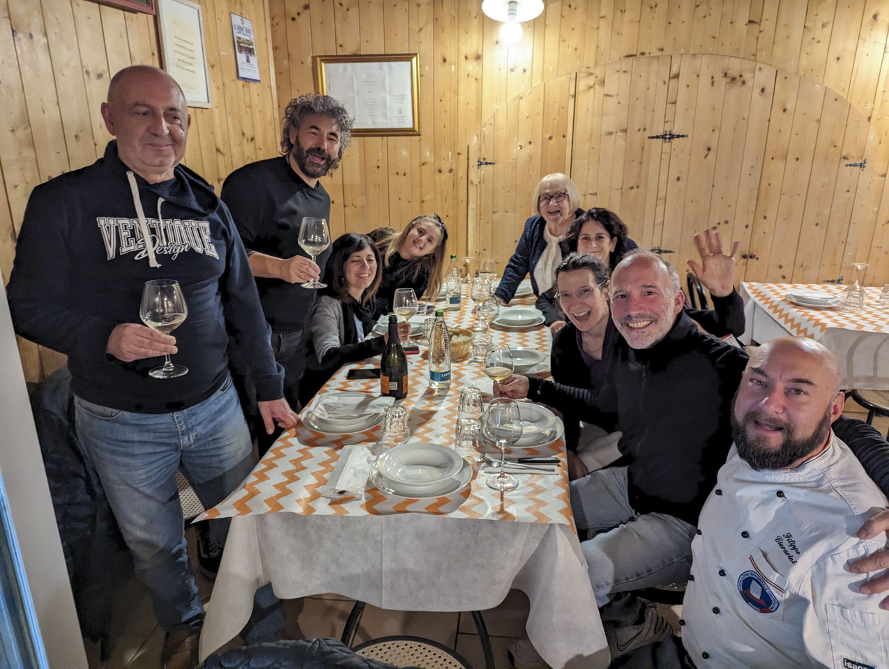 People gathered around a table and looking at the photo taker. There are glasses with some white wine and white empty dishes on the table. The room has light wood walls.