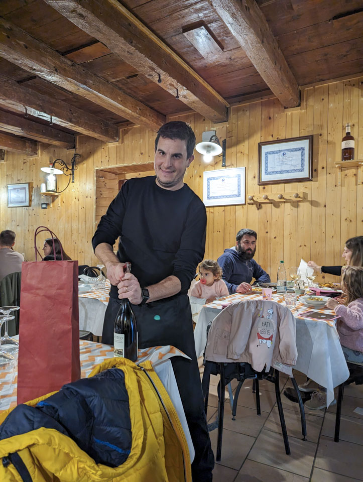 A man is opening a bottle of wine. Closer to use there is a tall red bag on the table and a yellow jacket on the back of the chair. There are three tables behind him, and customers are at two of them.