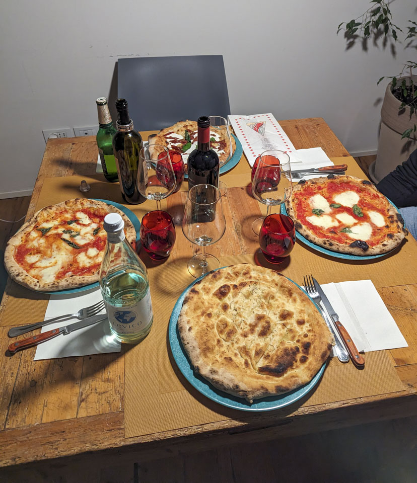 Four pizzas on the table. The one closest is just crust with rosemary. The two on the sides have cheese and basil, and the far one has other toppings.