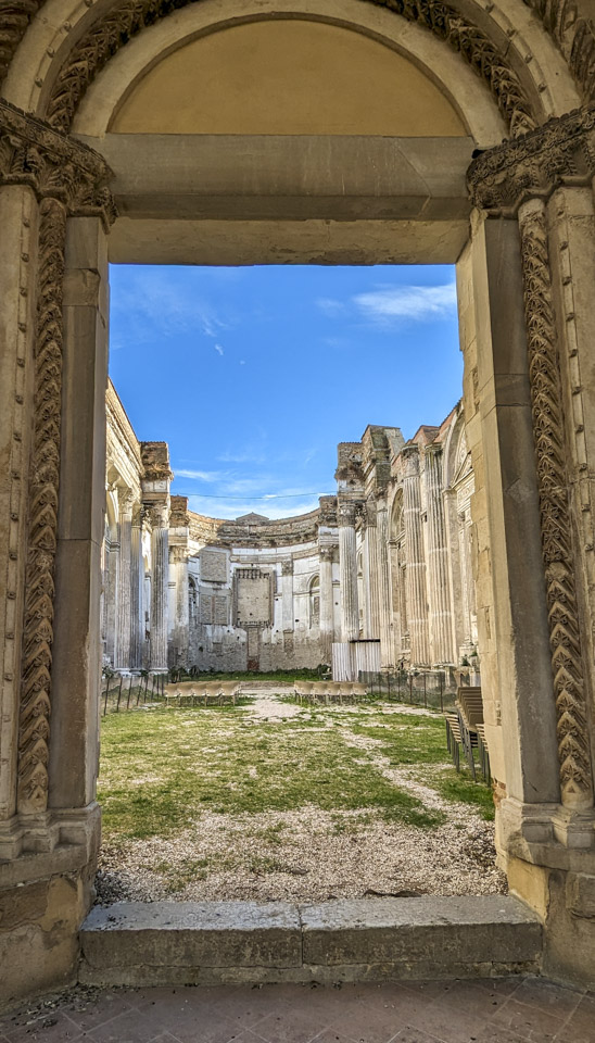 Looking through an empty doorway into a large space with columned walls on the sides but no roof. There are some rows of chairs in the center, plus some additional chairs stacked along one side.