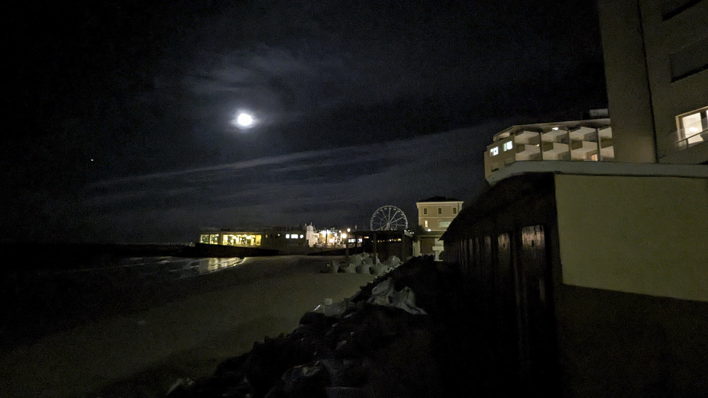 A view of the seaside in Pesaro, with a Ferris wheel visible, buildings with lights, and the moon obscured by clouds.