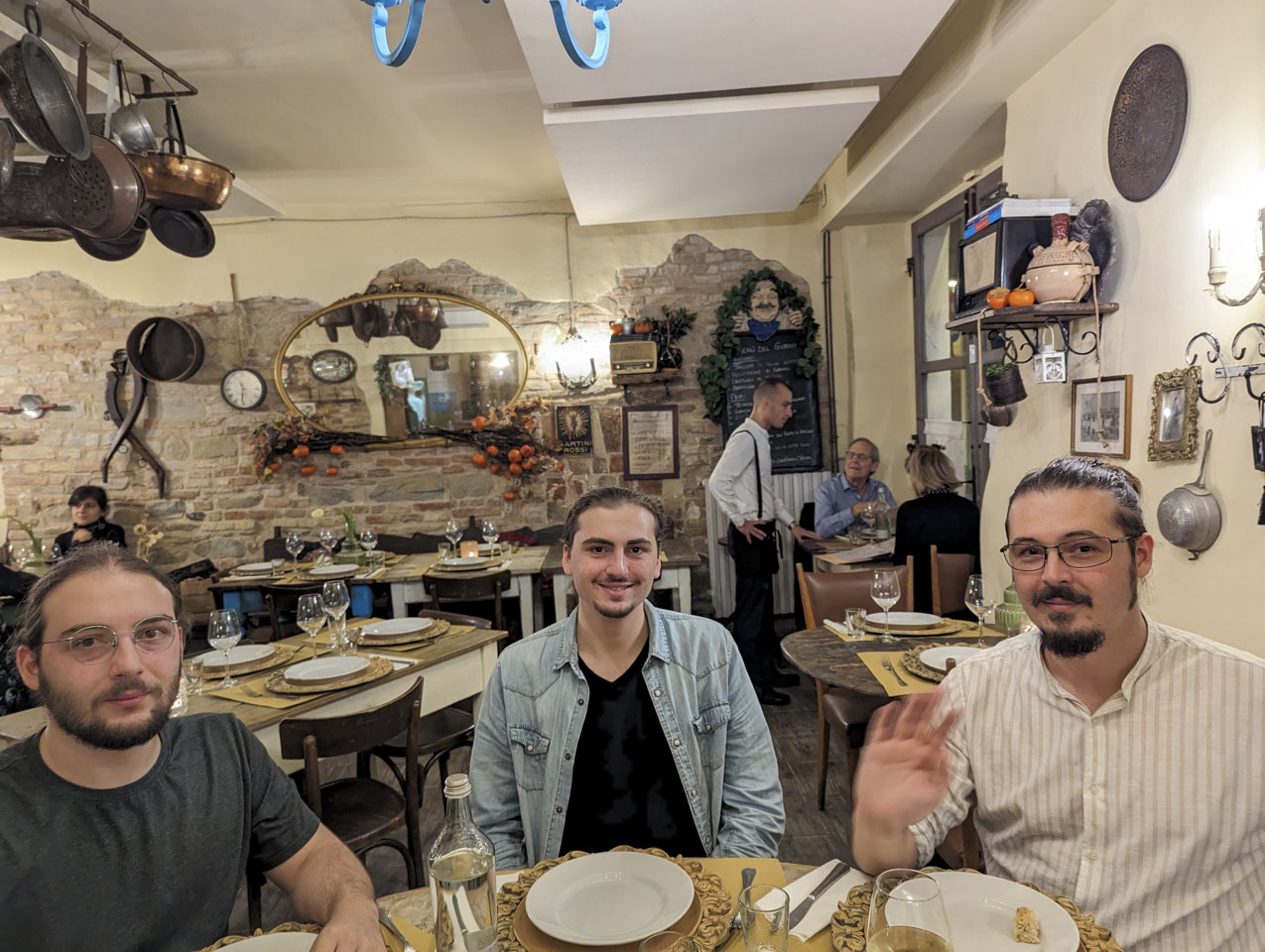 Three young men sitting at a restaurant table. In the background a waiter is at a table with two customers. The walls show some exposed old stones and some plaster.