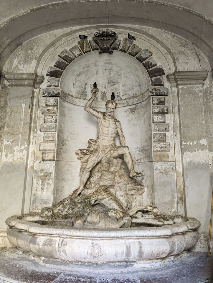 A stone or cement statue of a man with a raised arm, sitting on rocks, looking down at a creature coming out of the fountain below.