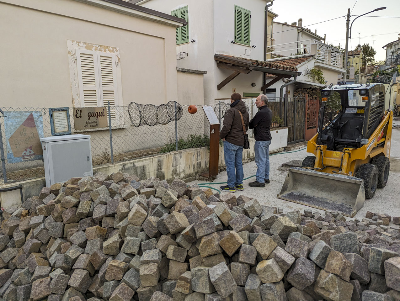 Francesco and Paul are reading a sight while standing next to a small front-loader. There is a pile of stones in the foreground. A sign that says El Gugul in on a fence.