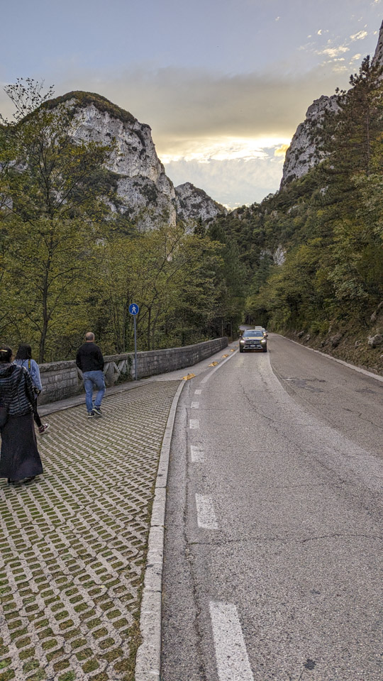 Tall hills have a road going between them, with some cars coming towards you. On the left is a patterned stone area with Anne, Flo, and Francesco walking away from you.