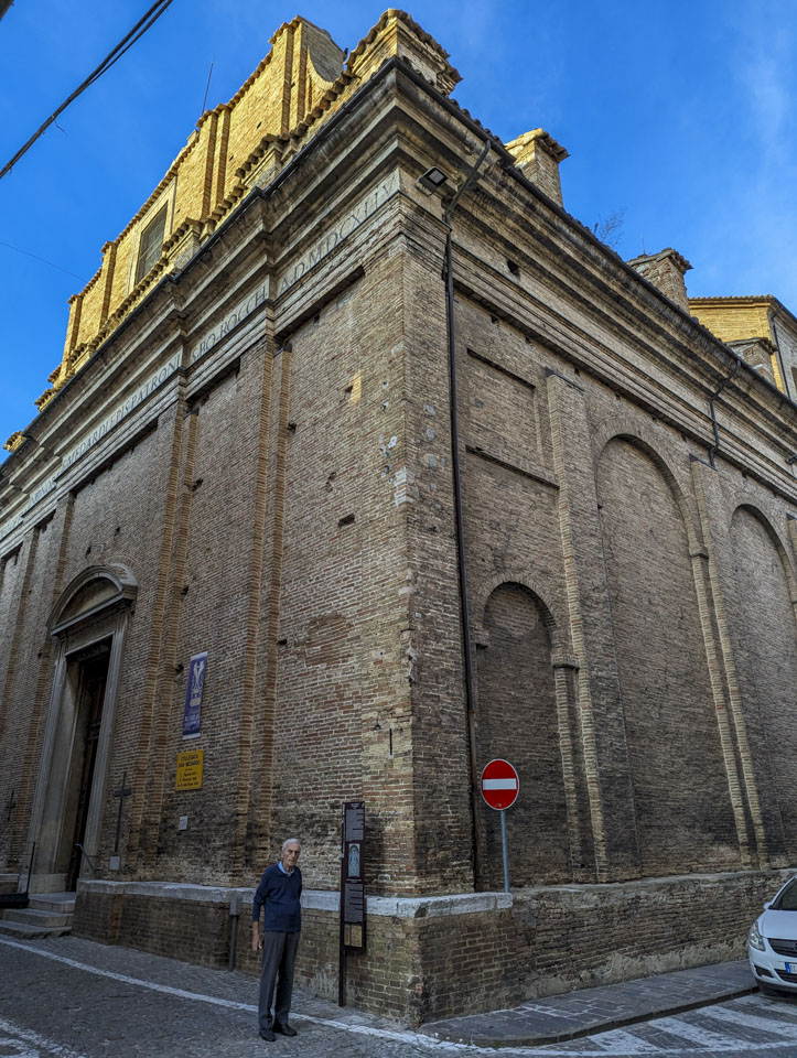 A thin elderly man in front of a large stone building