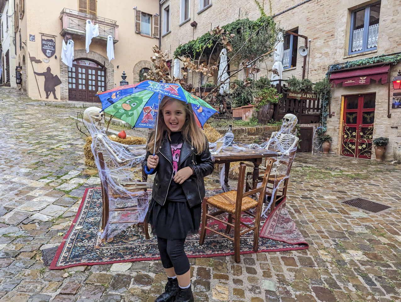 Gulia holding an umbrella while standing in front of a table that has a skeleton at each end, cards and a liquor bottle on the table, and fake spider webs all over everything.