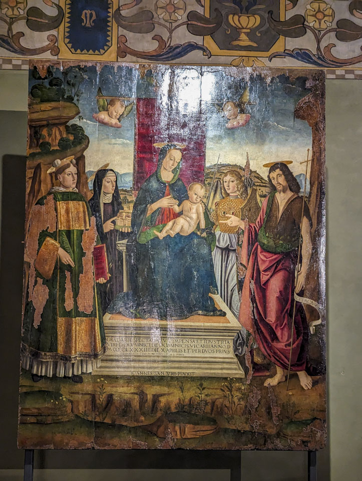 Painting of Mary wearing traditional blue and sitting on a throne while holding her child. Two people are standing on each side of her. The painting has damage, on the left side.