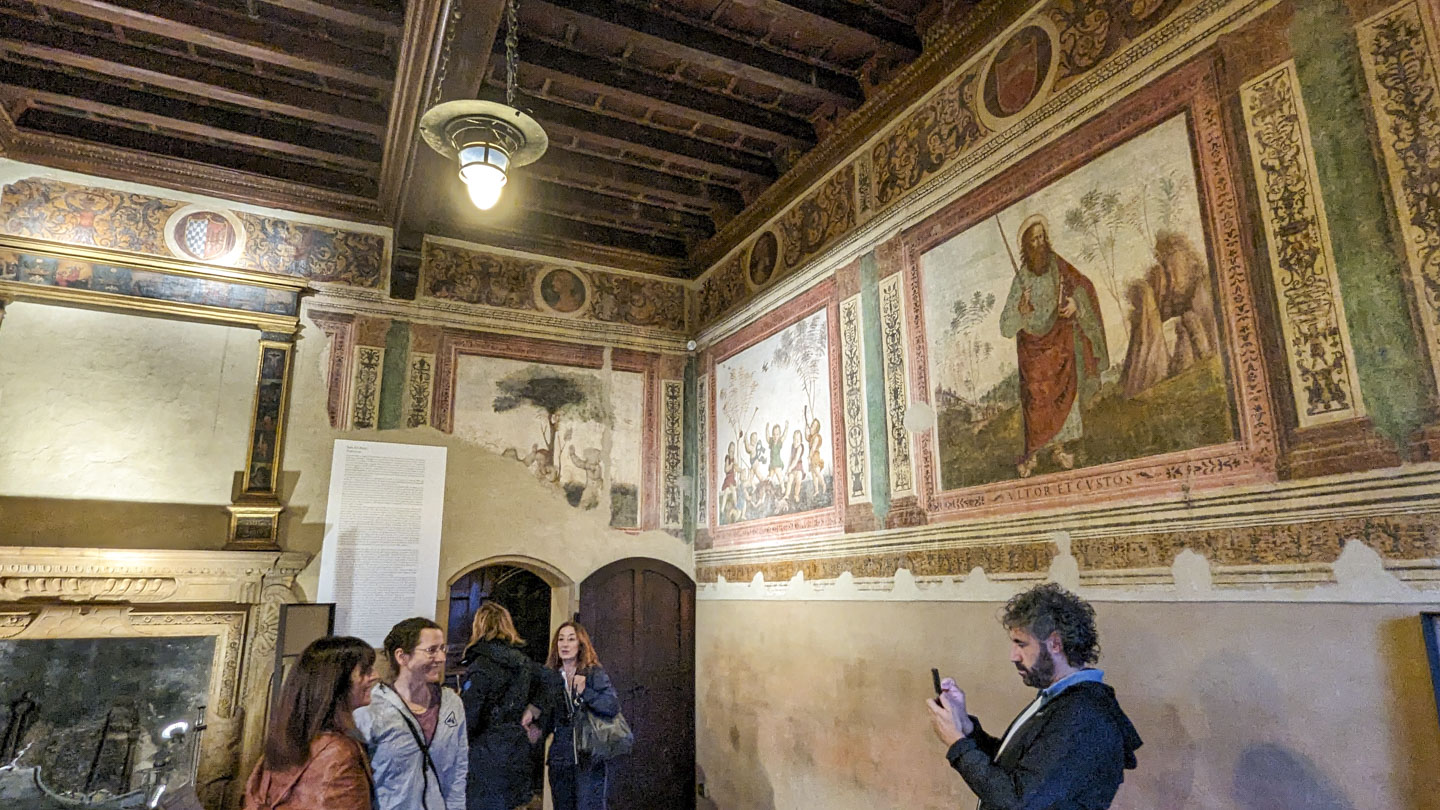 Federico taking a picture of Valentina and Anne while standing in the Putti Room in the Gradara Castle. A couple other tourists are also in the photo. The decorations on one wall are in good shape, but on the second wall they are damaged.