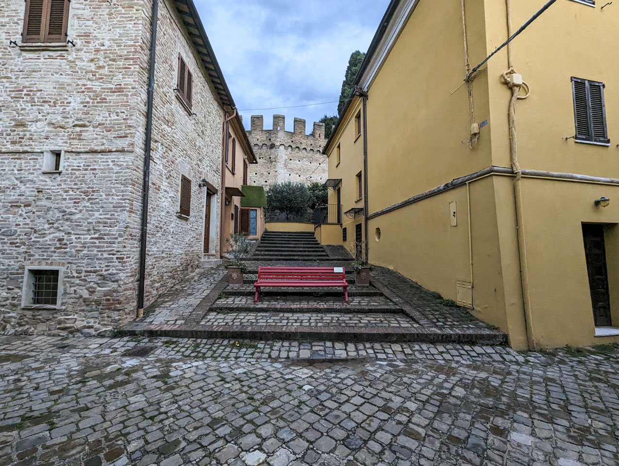 A red bench in an area between two buildings, with the wall of Gradara Castle seen in the background