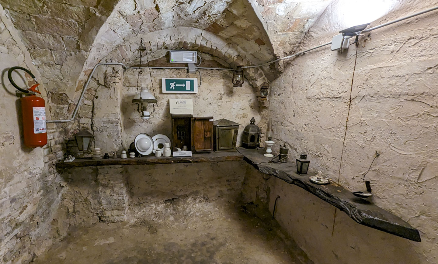 A small underground room with a shelf along 2 of the 3 visible walls. On the shelf are various lanterns and candles. There is also a fire extinguisher, a sign indicating the way to an emergency exit, and a bright electric light that is pointed at the ceiling.