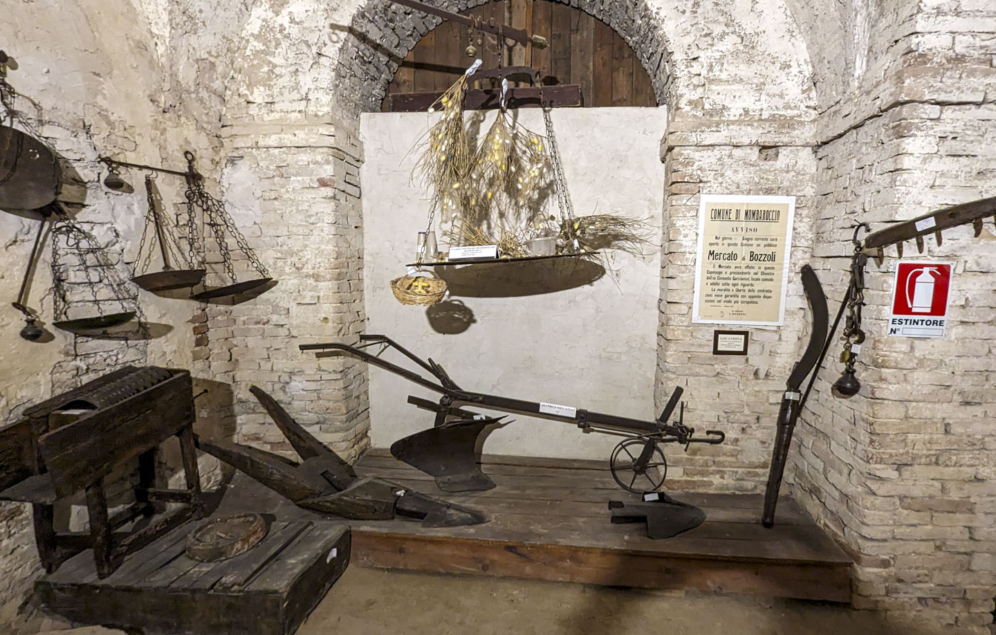 In the center of the photo is a large platform of a scale with some sample mulberry bush branches and silk worm cocoons on it. Below that is a plow. On the left are more scales. 