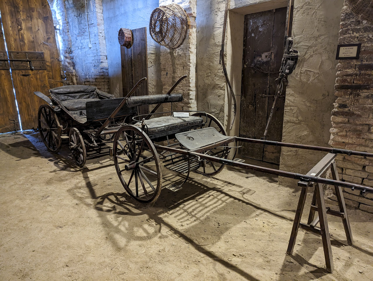 Two old black carriages, or buggies, one having only two wheels and the other having 4, both with long poles to secure to horses.
