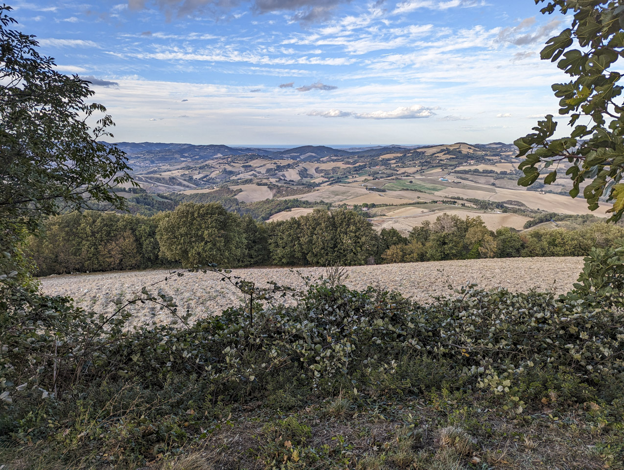 A blue sky with many white clouds has an agricultural landscape below, with tilled fields and high hills, while in the distance the flat Adriatic Sea can be seen.