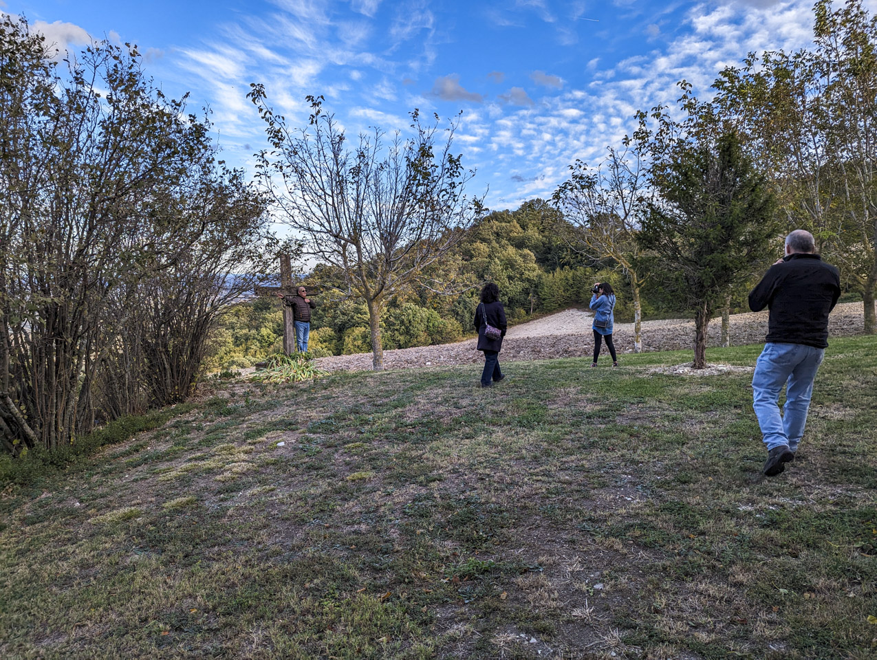 Francesco against a wooden cross, Doni walking towards Flo who is taking a picture, and Paul is getting ready to take a picture with his smartphone. The blue sky is above the late autumn landscape.