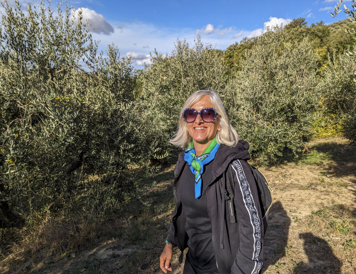 Alizia is wearing large sunglasses, black athletic clothing, and a neck scarf. Behind her are olive trees that are not too much taller than her.