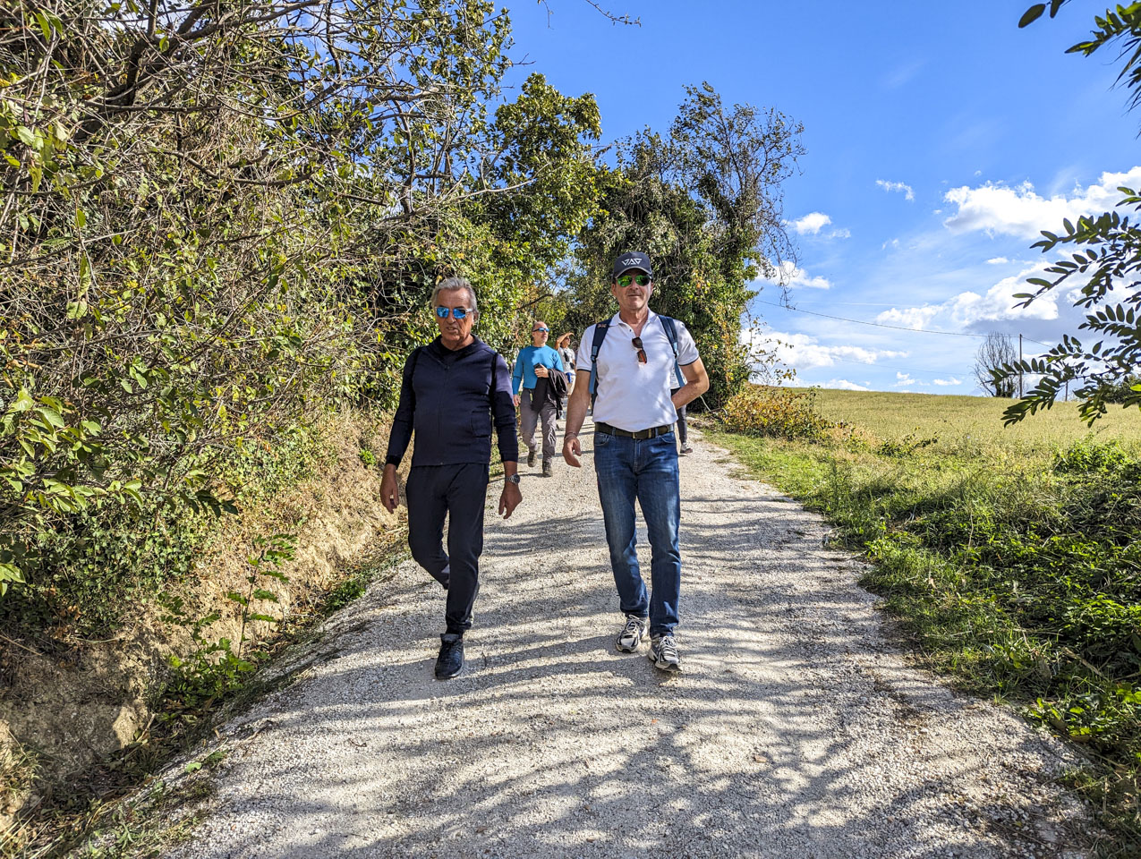 Two men walking down a white gravel road. Paul can be seen behind them. There is a field on the right and bushes on the left. It is a sunny day.