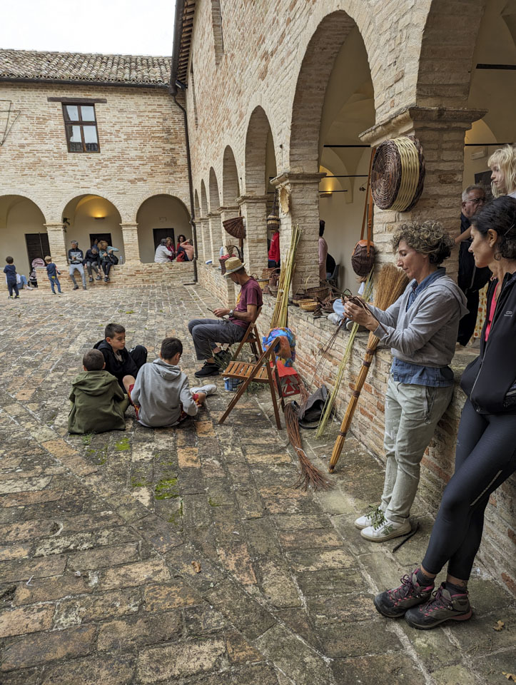 A courtyard with two walls of the stone building visible. A couple women are leaning against the wall; one of them is weaving. There are a number of other people visible as well.