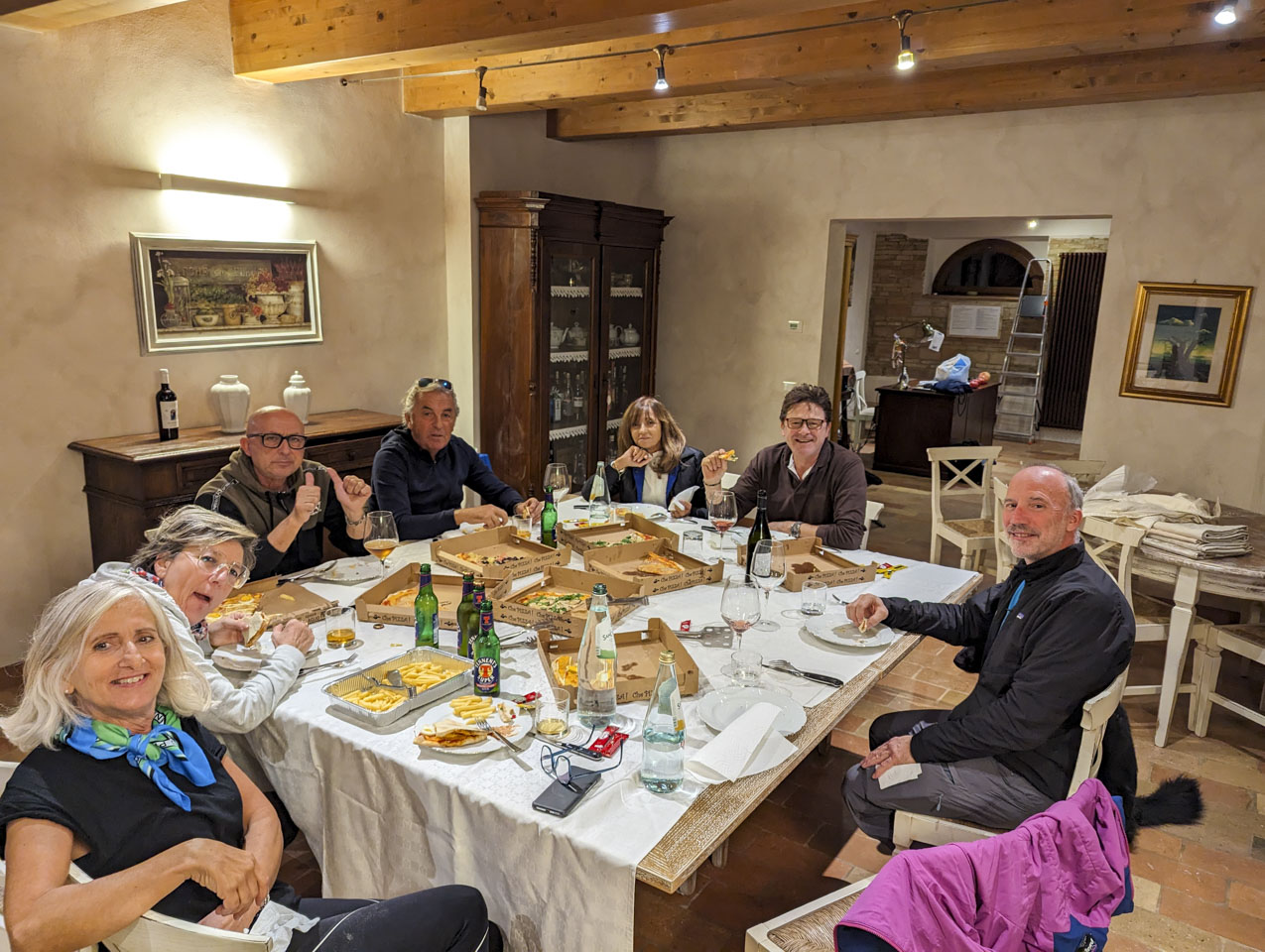 7 people sitting around the table with one empty chair. Pizza boxes with some pizza left, empty beverage bottles, and glasses on are the table. There are a couple paintings on the wall, sturdy wood beams along the ceiling, and a cabinet with dishes and bottles in it. Through an open entry can be seen another large room.