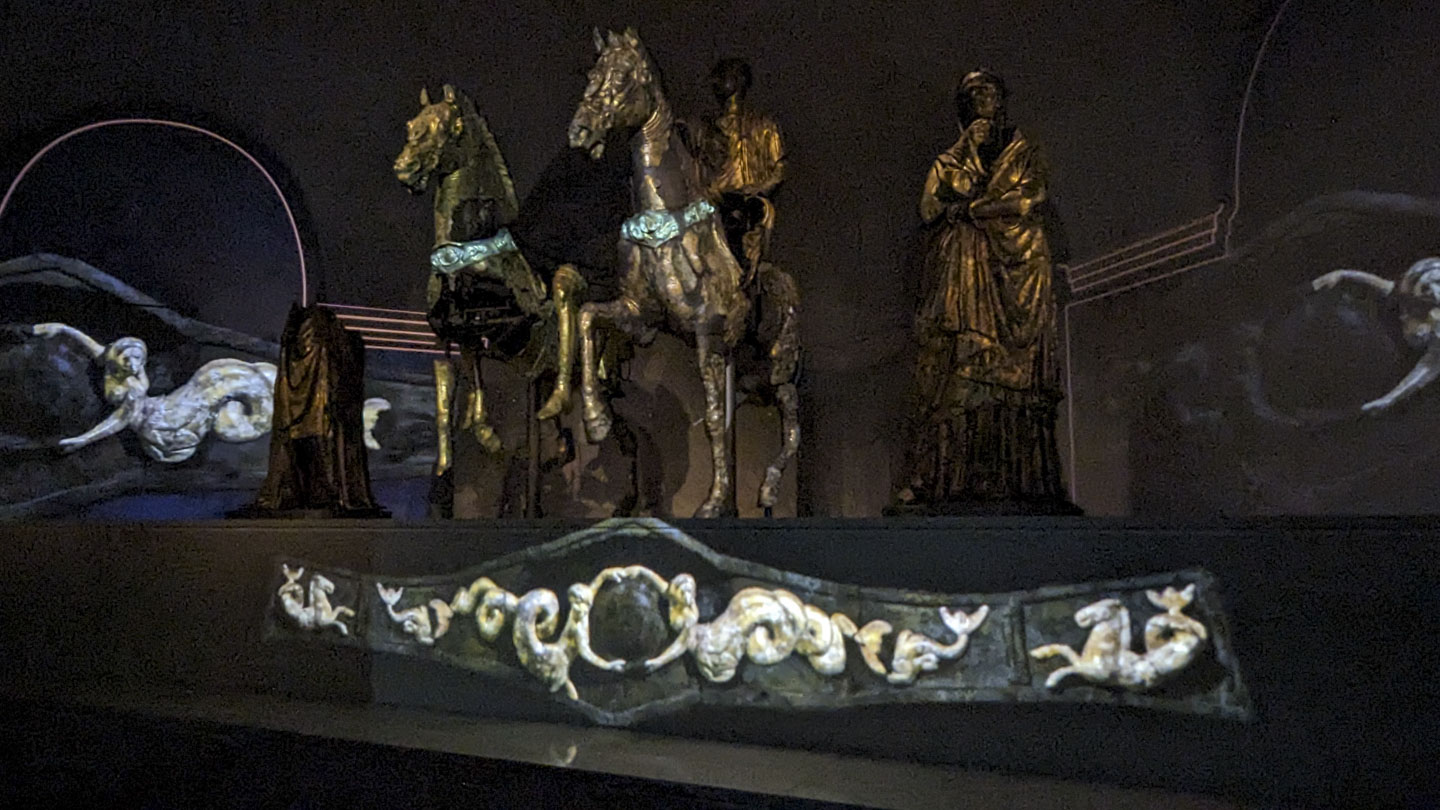 A picture of a darkened room with the ornamentation on the horses' chests illuminated in green. There is also an image of the chest plate shown on the wall.