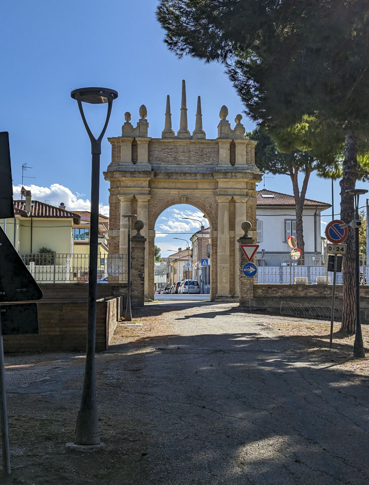 Looking down a path through a stone archway and then down a street. The sky is blue with a few clouds; there is a lamppost on the side of the path.