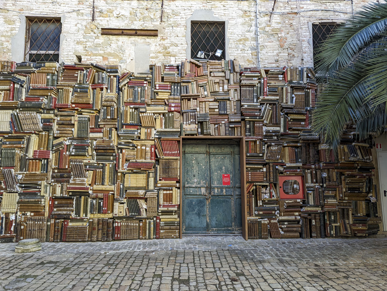 An outside wall with large books arranged in alternating directions like bricks are stacked. There is a plain green door in the middle of the wall.