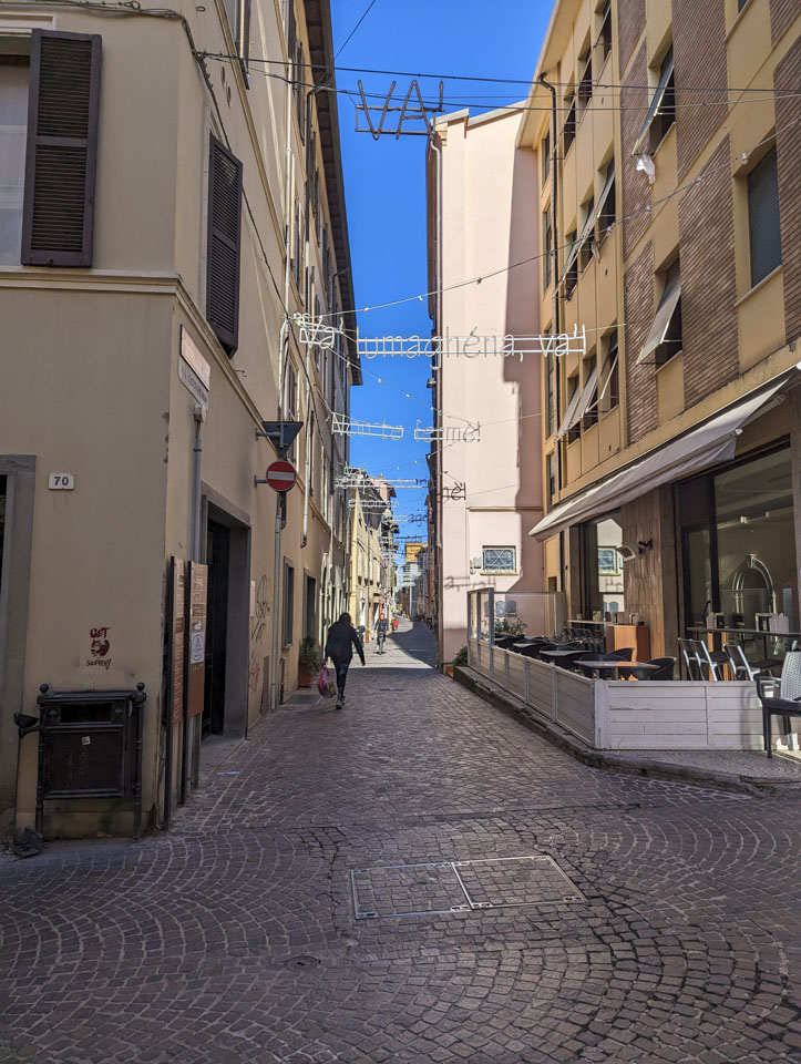 A brick road goes down a narrow street with 4 story buildings on both sides. Between the buildings are words that can be illuminated at night.
