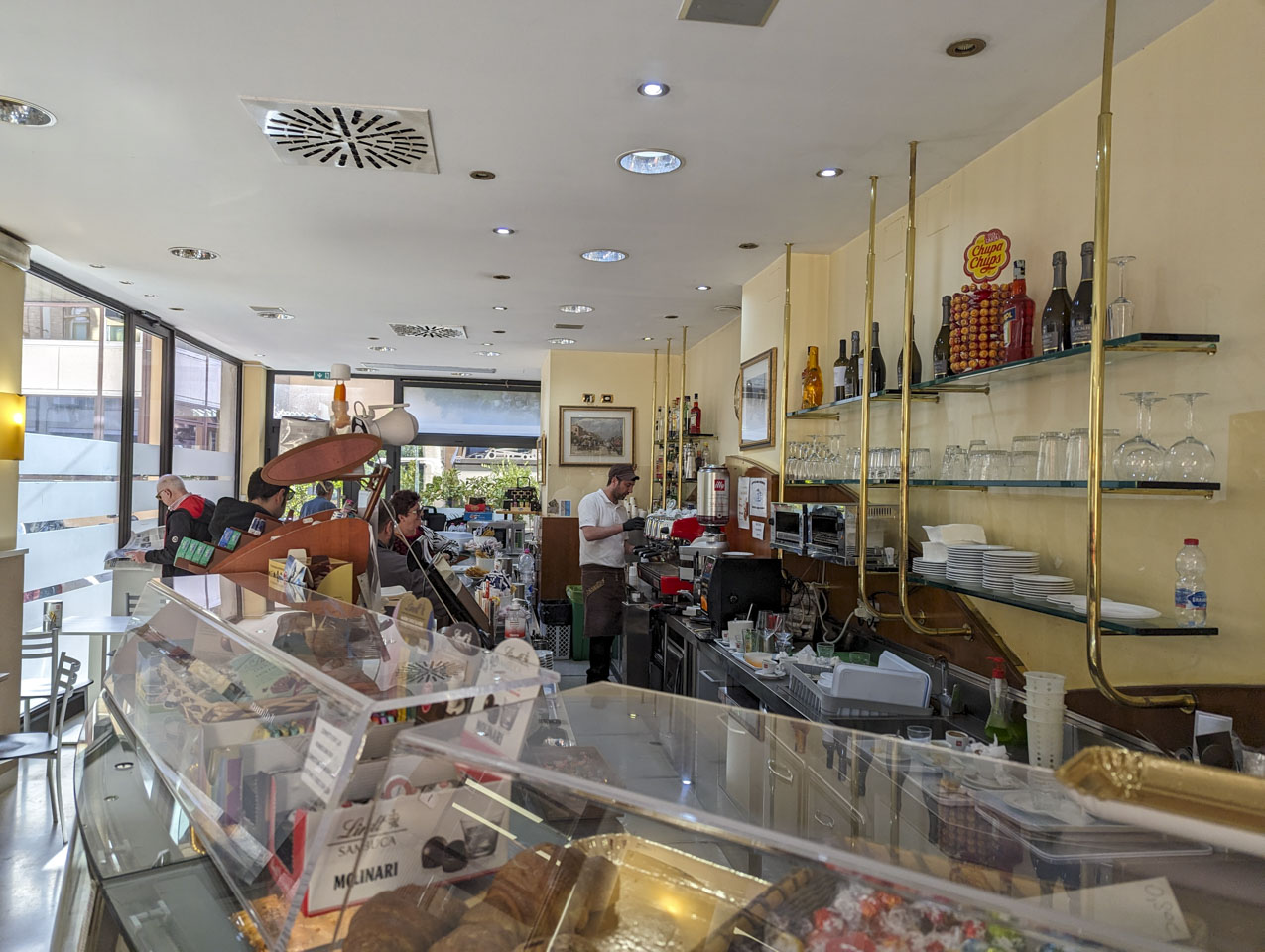 A pastry shop display counter with a man making a coffee drink behind it and some customers waiting.