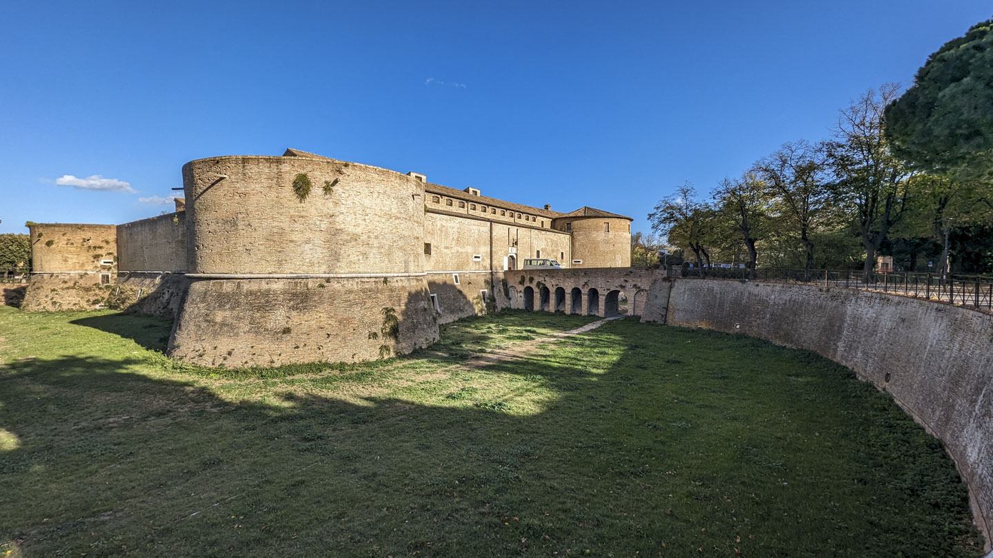 Looking from a wall across a recessed area to a strong stone building with round towers at each corner.