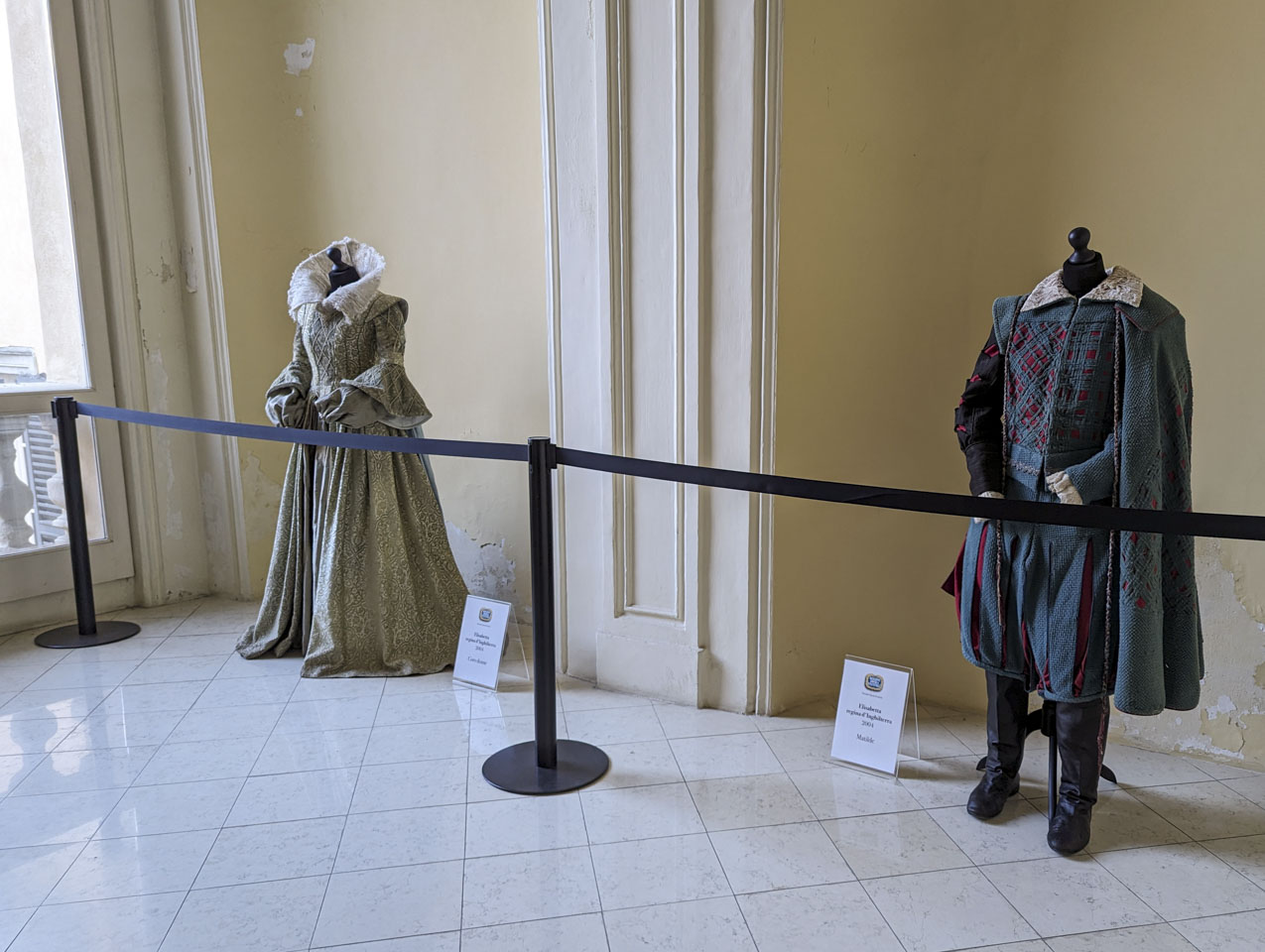 On a large landing area with a tile floor, behind a rope is an elaborate Elizabethan dress and on the right is a courtly outfit with pants and a cape.