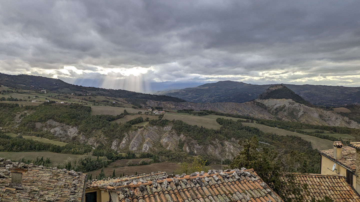 Rays of light stream from an opening in the clouds and land behind the ridge of a mountainside. There are a few peaks visible. A few rooftops can be seen in the foreground at the bottom of the picture.