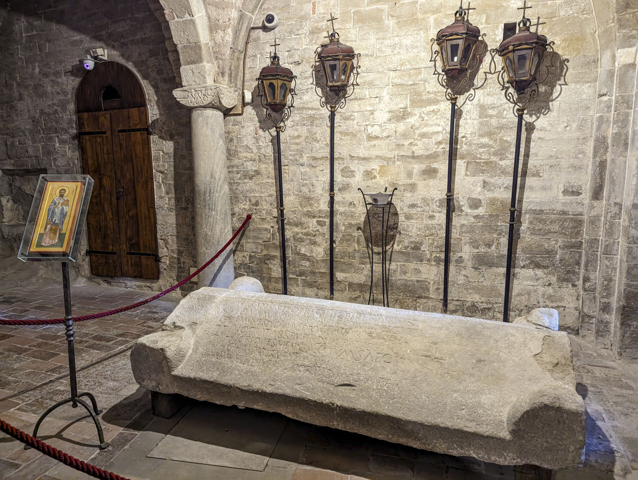 The lid to a tomb with a picture of San Leo on a stand near it. There is some writing inscribed into the lid.