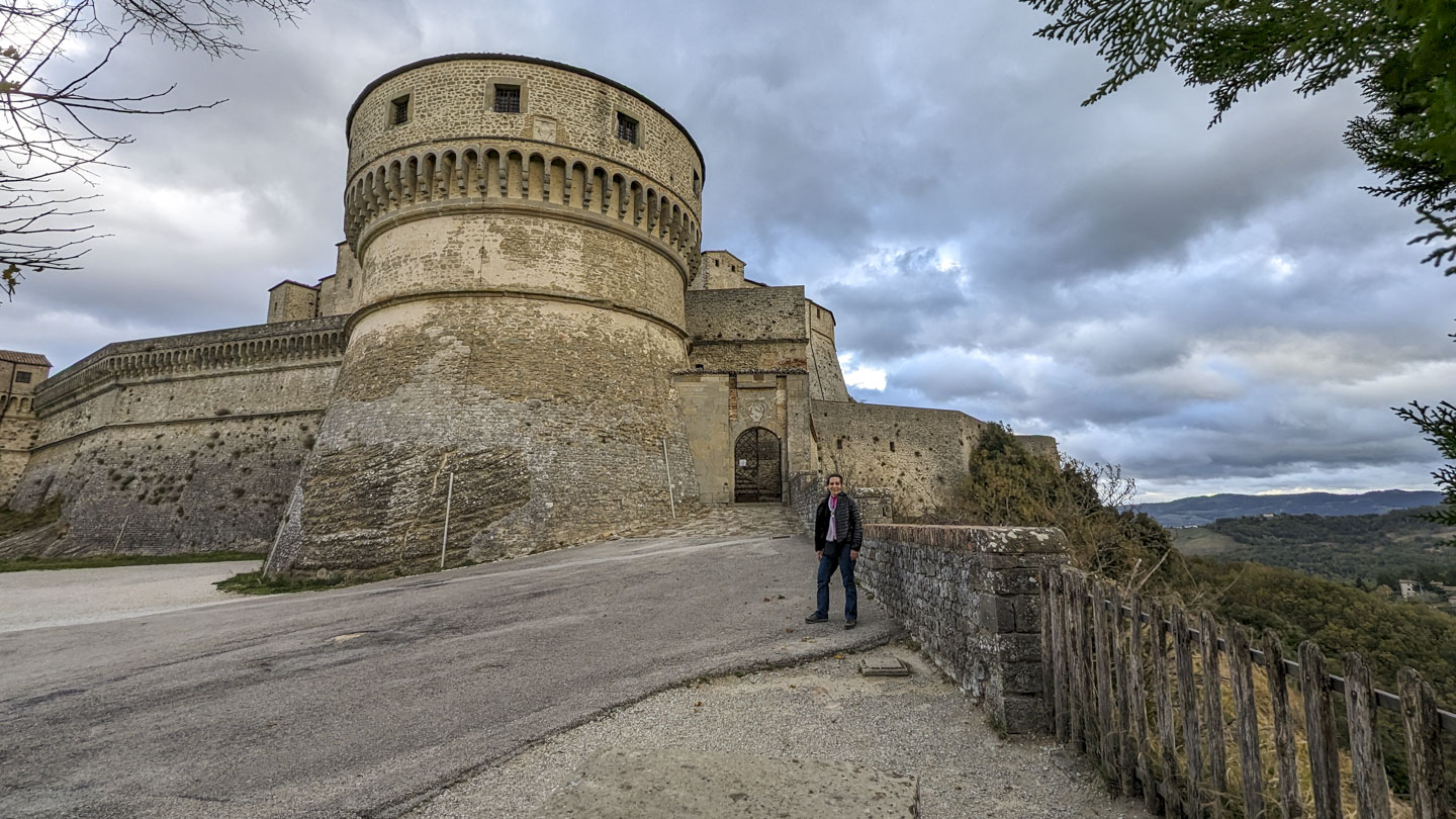 Anne is standing next to a short stone wall with the fortress behind her. A large tower is next to an entrance that has a grate door. The land falls off to the right, behind the short wall.